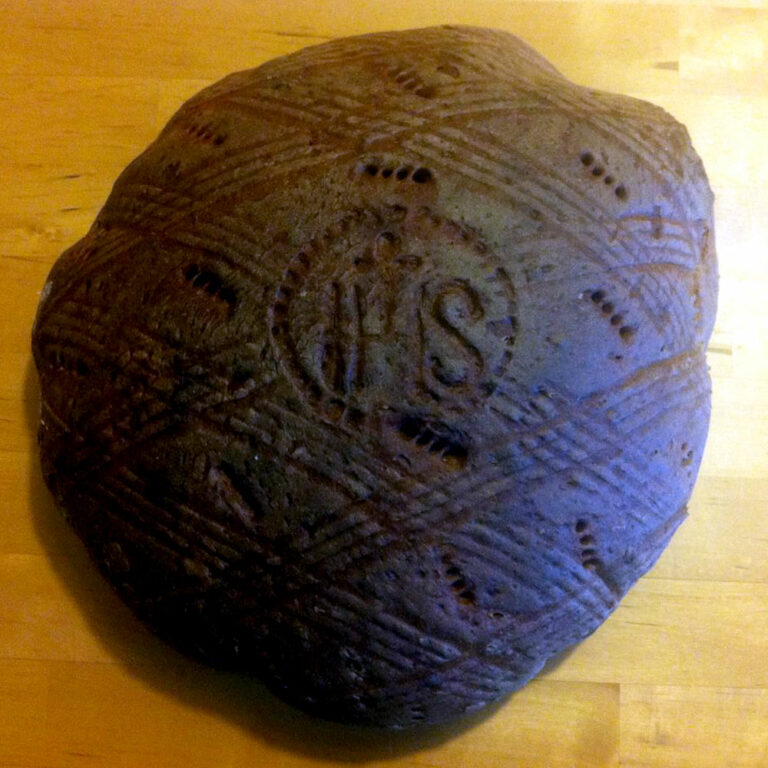 Ein runder Laib Roggenbrot mit komplizierten Mustern und kleinen Löchern in der Kruste. In der Mitte sind die Buchstaben „IHS“ eingeprägt. Das Brot wird auf eine Holzoberfläche gelegt.