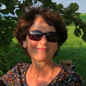 Eine Frau mit kurzen braunen Haaren und Sonnenbrille sitzt lächelnd draußen. Sie trägt ein gemustertes Oberteil und das Sonnenlicht wirft einen warmen Glanz auf ihr Gesicht. Hinter ihr ist ein kleiner Hund teilweise sichtbar und grünes Laub umrahmt die Szene.