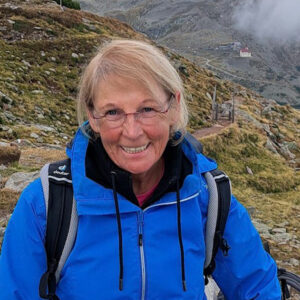 Eine Person in blauer Jacke und Rucksack lächelt beim Wandern auf einem Bergpfad. Die Landschaft besteht aus Grasland und in der Ferne aus felsigen Hügeln unter einem bewölkten Himmel.