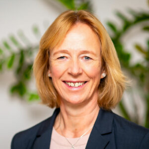 Eine Frau mit schulterlangem blondem Haar lächelt warmherzig. Sie trägt einen schwarzen Blazer über einem hellen Oberteil. Im Hintergrund ist eine verschwommene grüne Pflanze zu sehen, die eine Inneneinrichtung suggeriert.