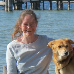 Eine Frau mit Brille lächelt, während sie neben einem braunen mittelgroßen Hund steht. Sie sind draußen in der Nähe eines Gewässers mit einem Holzsteg im Hintergrund. Die Frau trägt einen leichten Pullover und der Hund hat ein Halsband.