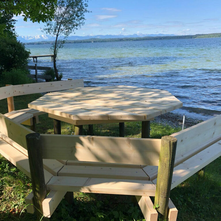 Ein achteckiger Holztisch mit Bänken steht auf üppigem Gras neben einem See. Das Wasser plätschert sanft unter einem klaren blauen Himmel, in der Ferne sind Berge zu sehen. Bäume spenden teilweise Schatten über dem Sitzbereich.