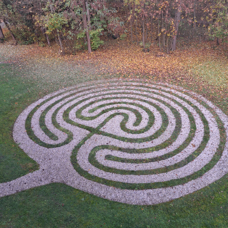 Luftaufnahme eines kreisförmigen Kieslabyrinths mit einem einzigen gewundenen Pfad, der von grünem Gras und Herbstbäumen umgeben ist. Die Blätter an den Bäumen zeigen Schattierungen von Gelb, Orange und Braun, und einige abgefallene Blätter liegen verstreut auf dem Boden.