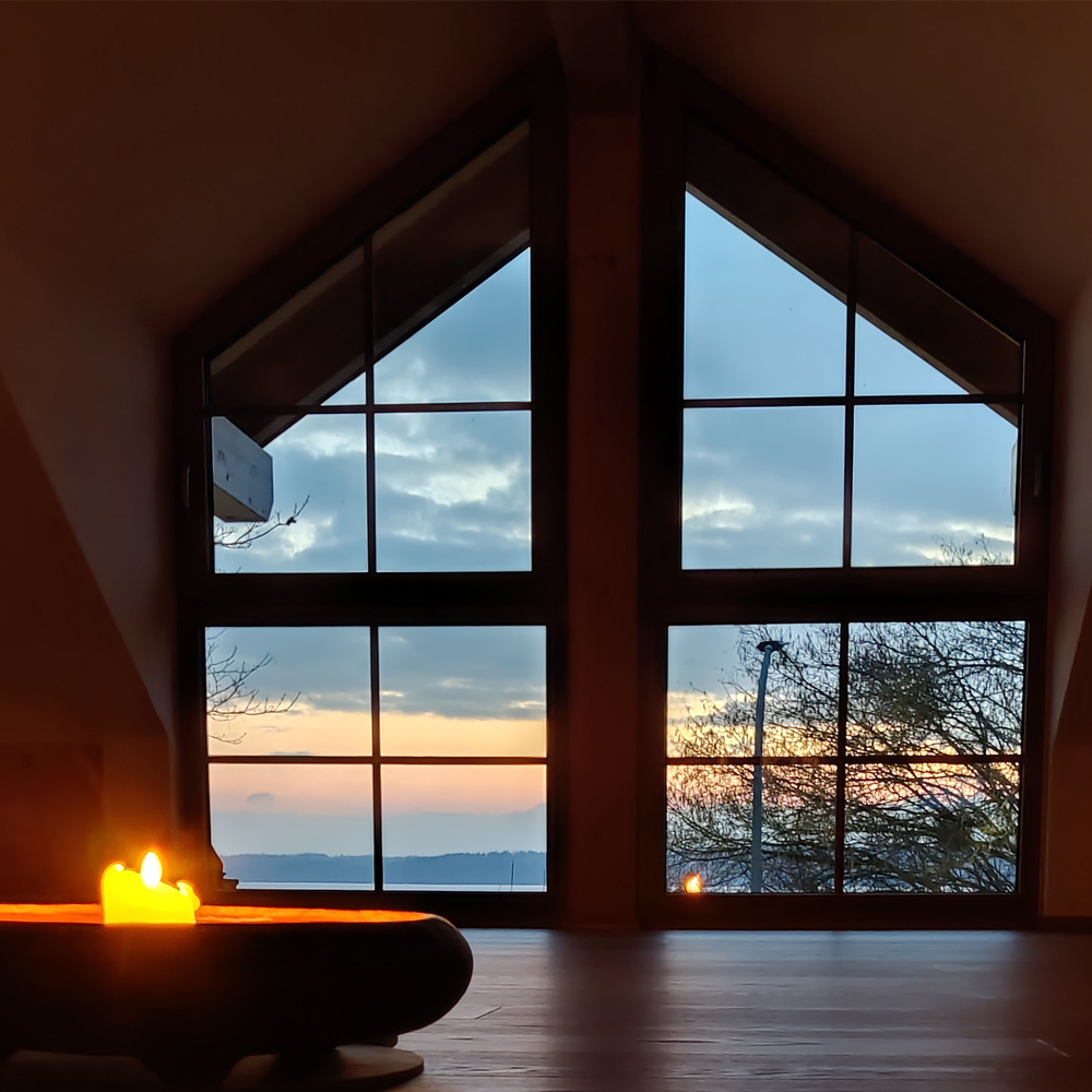 Ein schwach beleuchteter Raum mit großen dreieckigen Fenstern, die einen Blick auf einen Sonnenuntergang bieten. Im Vordergrund stehen zwei brennende Kerzen, die ein warmes Licht verbreiten. Draußen sind Baumsilhouetten und ein teilweise bewölkter Himmel sichtbar.
